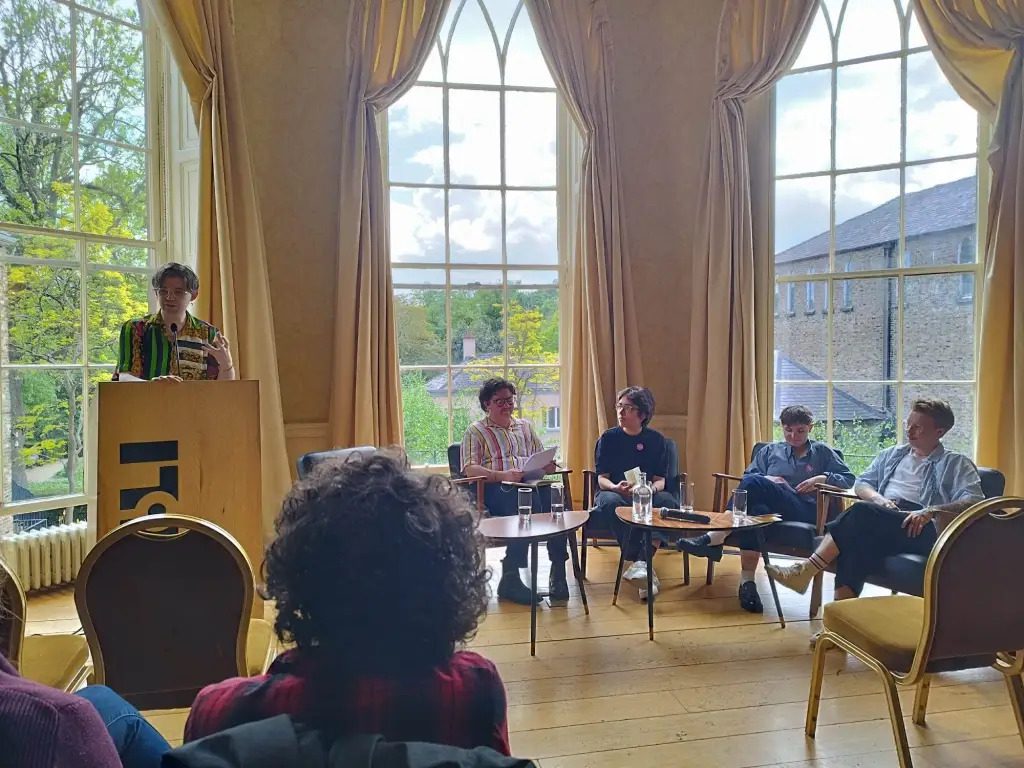 Photo in a well-lit room showing five people. One person is standing at a podium, the rest are seated around two small tables.