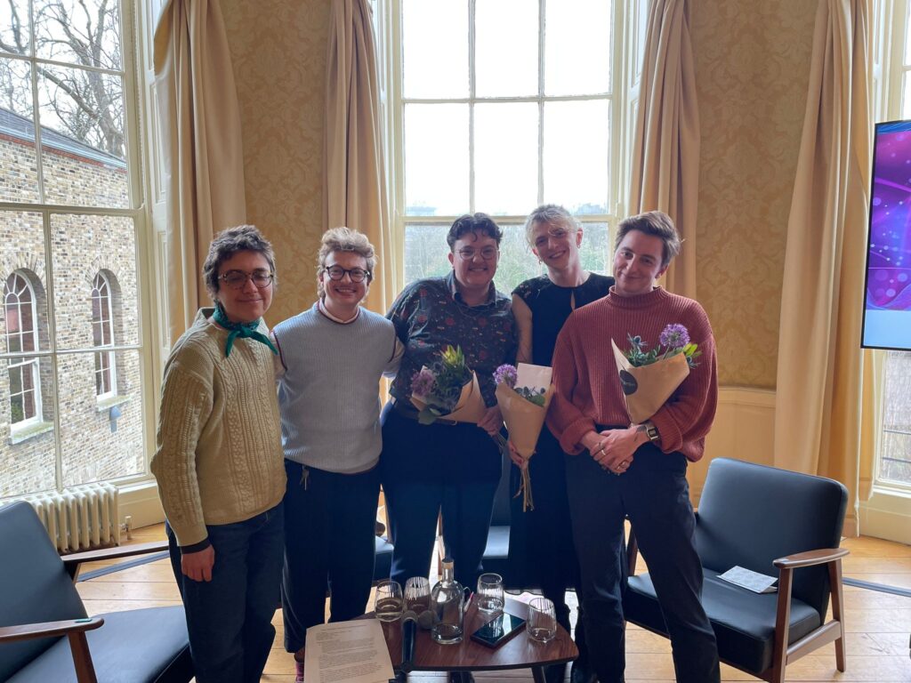 Five individuals stand in the Physics theatre in MoLI. They are, left to right, Robin Steve, a nonbinary person with glasses and short brown hair, and a cream jumper; Matt Kennedy, a trans man with blonde hair and glasses and a blue sweater vest; Taylor Follett, a trans man with brown hair and glasses wearing a button-down shirt and holding flowers; Alexa Moore, a trans woman with blode hair pulled back wearing a black blouse and skirt and holding flowers; and Daire Dempsey, a nonbinary [erson with short brown hair and a orangey-red jumper holding flowers.