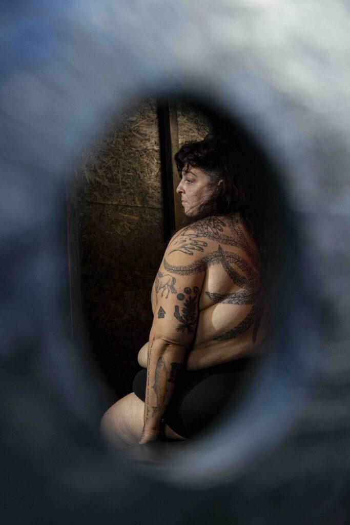 A person with tattoos and dark hair faces left and is profiled. They are topless and have a number of intricate tattoos. They are in the centre of a blurred frame